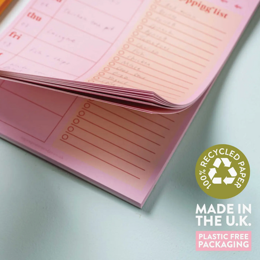 Pink notebook with a recycle logo and text about recycled paper and plastic-free packaging on a light gray background.