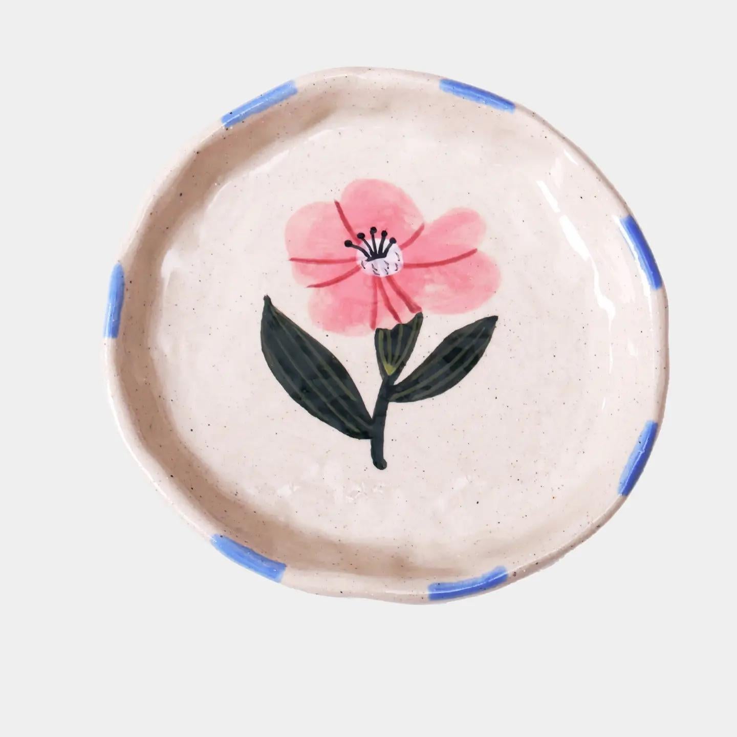 βHand-painted stoneware plate with pink flower design and blue rim brushstrokes on a white background.β
βClose-up of small ceramic dish with pink floral design used to hold gold jewelry.β
βDelicate pink flower plate displayed on a pastel table with dried petals and a cup of tea.