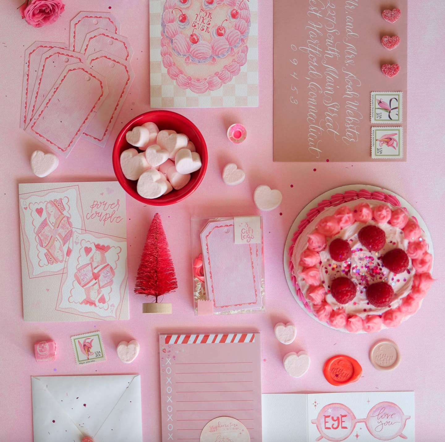 Pink-themed stationery items including cards, envelopes, and a small bowl of pink candies on a pink background.