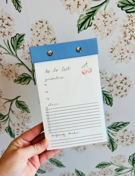 “Cherry Bow Notepad” – Notepad with cherry print and blue ribbon design styled with colorful pens

“Cute On-the-Go Notes” – Notepad peeking out of a tote with lip balm and planner accessories
