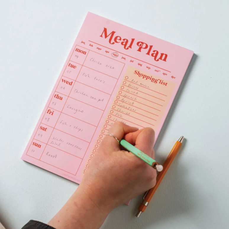 Pink meal plan and shopping list notepad with a hand holding a pen on a white surface