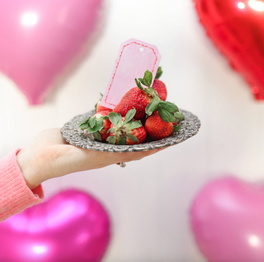 Hand holding a small dish of strawberries with pink and red balloons in the background
