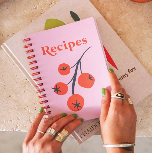 Pink recipe book with tomato illustrations held by a person on a light surface