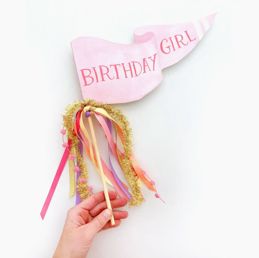 Pink birthday hat with 'Birthday Girl' text and colorful ribbons held by a hand on a white background