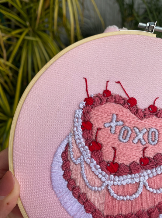 Embroidery hoop with a heart design featuring cherries and 'xoxo' text on a pink background.