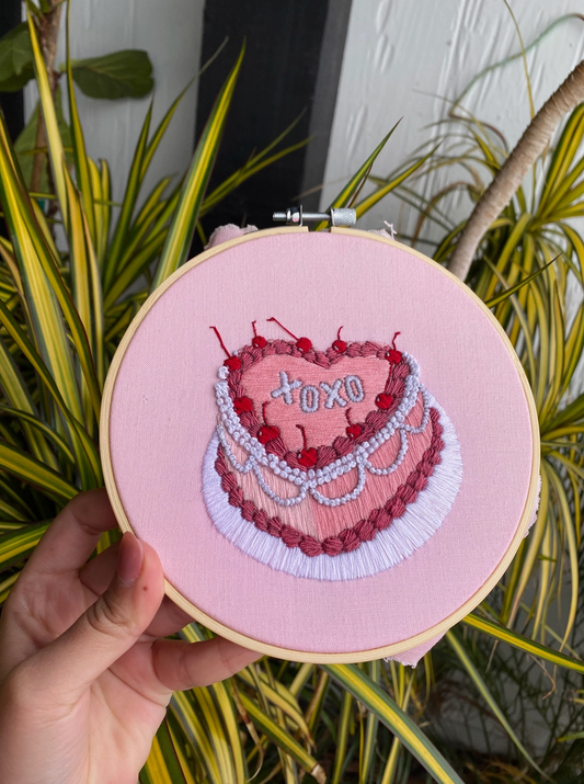Embroidery hoop with a pink cake design and 'XOXO' text, held against a plant background.